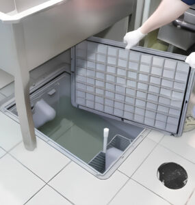 Technician in gloves lifting the lid off an in-floor grease interceptor in a commercial restaurant kitchen