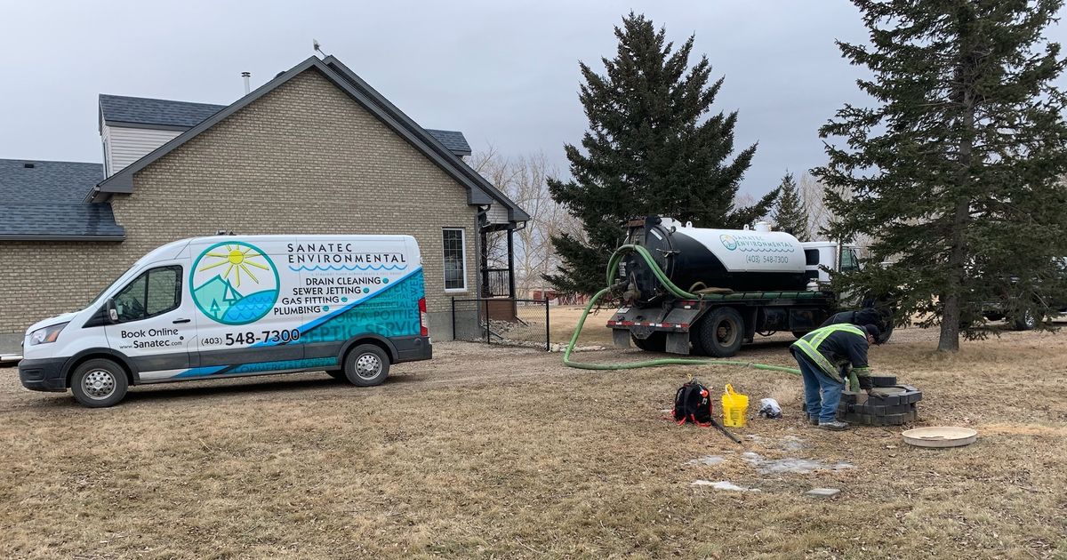 Sanatec Environmental vacuum truck with its hose run to an open septic tank on a rural acreage, a technician working at the tank and a Sanatec service van parked beside the house